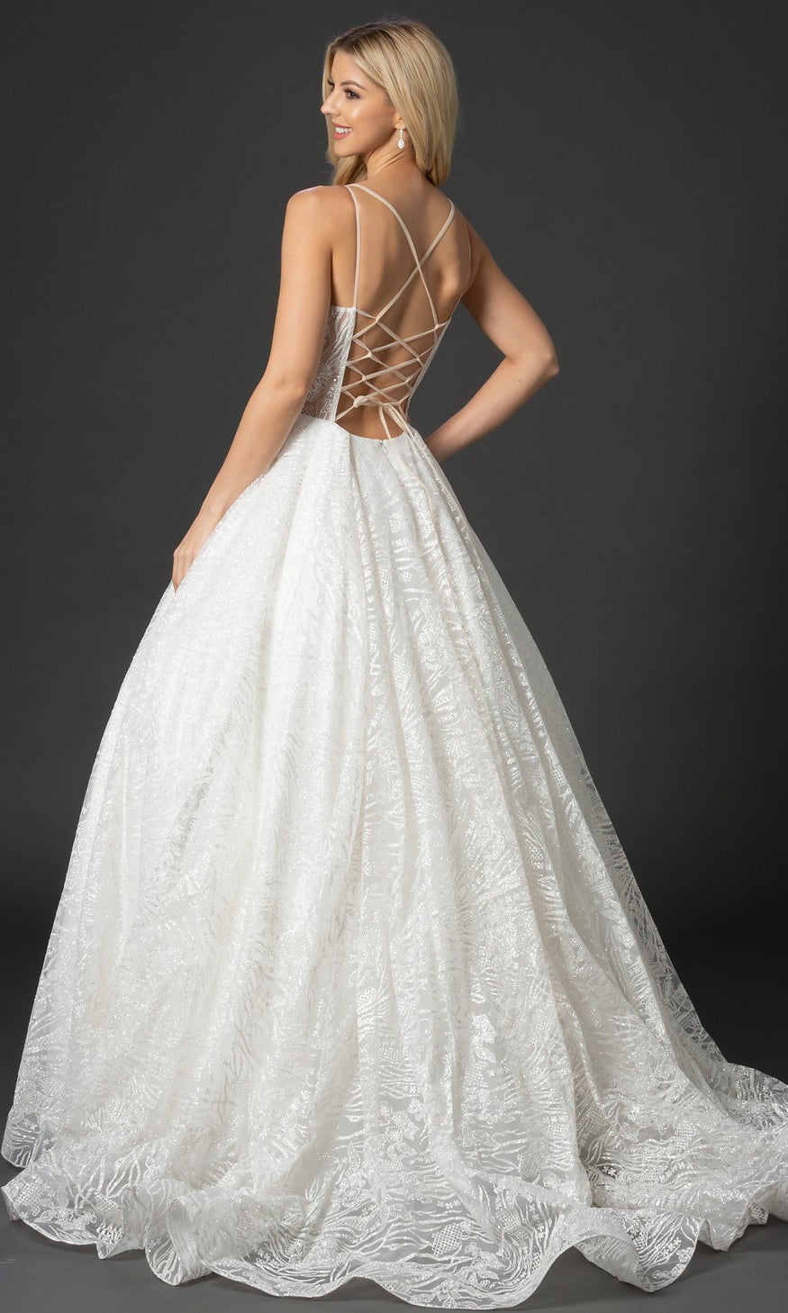 A woman in a white Nina Canacci 8202 Glitter-Print Ball Gown with a floor-length, sleeveless design and lace-up open back stands facing away, looking over her shoulder against a dark background.